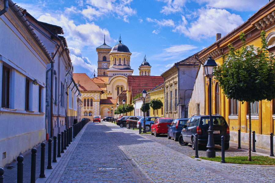 Alba Iulia Romania A narrow cobblestone street lined with colorful buildings and parked cars leads to a large, ornate church with domes and towers in the background. The sky is bright blue with scattered clouds, and a few small trees are on the sidewalk.