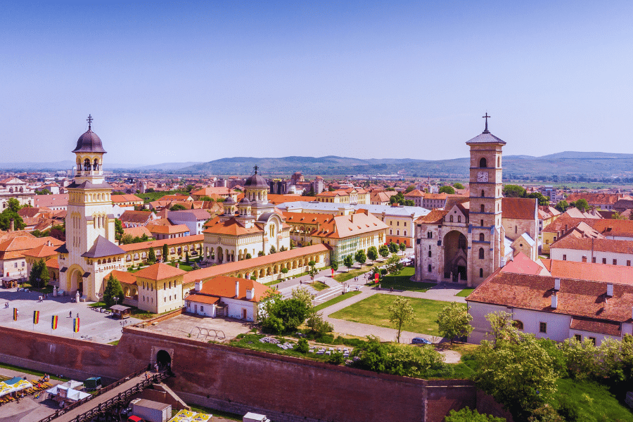 Alba Iulia Romania Aerial view of a historic European town featuring two prominent church buildings with imposing bell towers, surrounded by red-roofed houses. In the foreground, there is a fortified wall and lush greenery, with distant rolling hills under a clear blue sky.