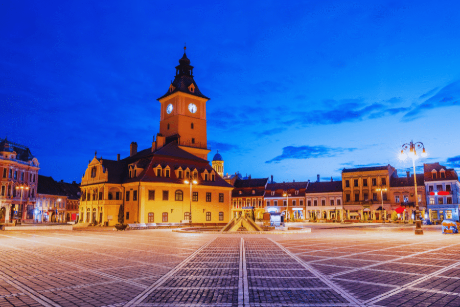 Brasov Romania Historical town square at dusk, featuring a large historic building with a clock tower, surrounded by well-lit buildings. The square's paved surface shines under street lamps, and the deep blue sky enhances the warm glow of the surroundings.