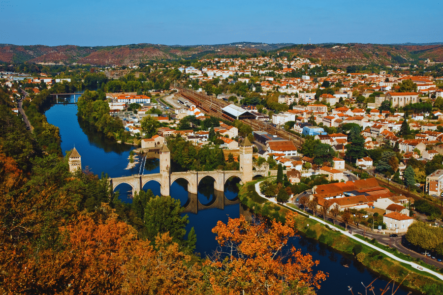 Cahors-France A picturesque aerial view of a medieval stone bridge with towers spanning a calm river, nestled in a vibrant town surrounded by rolling hills with autumn foliage. The town has a mix of modern and historic buildings, with clear blue skies above.