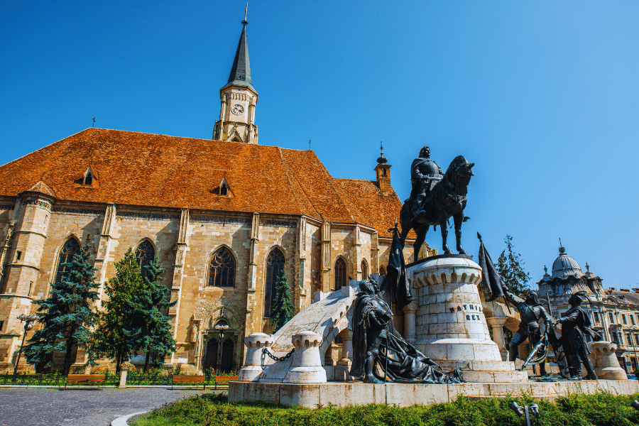Image of St. Michael's Church in Cluj-Napoca, Romania, with a tall clock tower and a reddish-orange roof. In the foreground stands a prominent statue featuring a mounted rider and several figures, set against a clear blue sky.