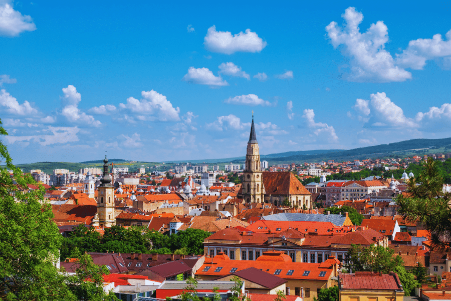 Cluj Napoca Romania A picturesque view of a city with vibrant red-roofed buildings, two prominent church towers, and lush green hills in the background. Fluffy white clouds dot the blue sky, enhancing the scenic landscape.