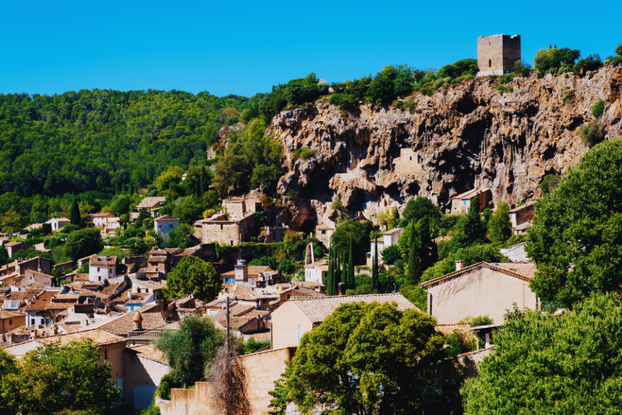 Cotignac France A picturesque village nestled at the base of a rugged, rocky cliff topped with a small, ancient stone tower. The village comprises quaint, tiled-roof houses surrounded by lush greenery and trees, all under a clear blue sky.