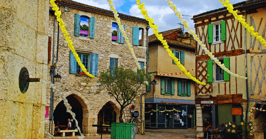 Eymet-Brittany-France A picturesque European village square with historic stone buildings, colorful shutters, and a small fountain to the left. Yellow streamers are strung across the square, adding a festive touch. Cafés and shops with outdoor seating line the charming streets.