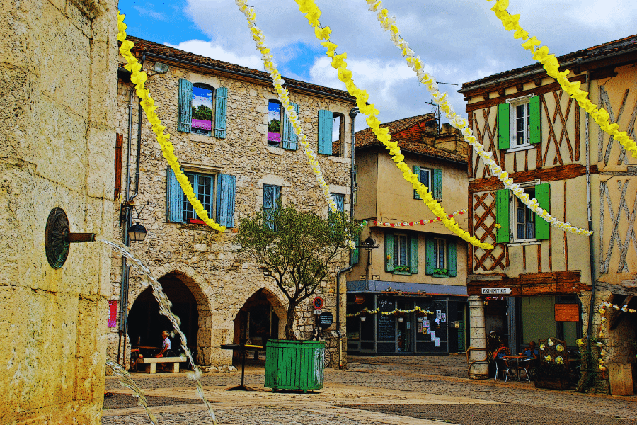 Eymet-Brittany-France A picturesque European village square with historic stone buildings, colorful shutters, and a small fountain to the left. Yellow streamers are strung across the square, adding a festive touch. Cafés and shops with outdoor seating line the charming streets.