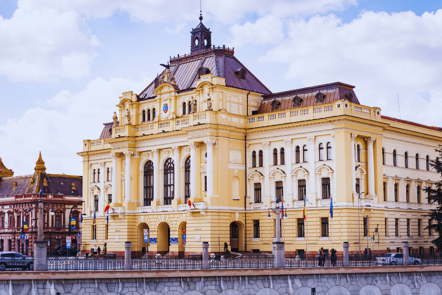Oradea Romania A grand, historic building with ornate architecture and a clock tower stands under a partly cloudy sky. The yellow and beige facade features arched windows and intricate details. People are gathered near the entrance, and flags are displayed outside.