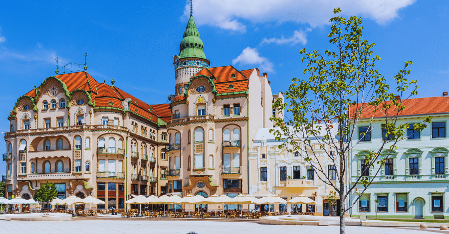 Oradea Romania A sunny view of a picturesque European town square with ornate, historic buildings featuring red-tiled roofs and a green turret. The square has outdoor cafes with umbrellas, a stone-paved ground, and a few trees, under a blue sky with scattered clouds.