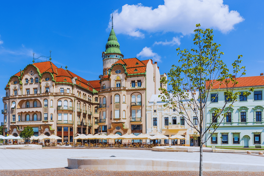 Oradea Romania A sunny view of a picturesque European town square with ornate, historic buildings featuring red-tiled roofs and a green turret. The square has outdoor cafes with umbrellas, a stone-paved ground, and a few trees, under a blue sky with scattered clouds.