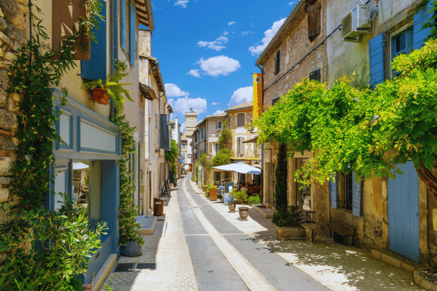 Saint-Remy-de-Provence-France A picturesque narrow street in a charming village, lined with rustic stone buildings adorned with pastel-colored shutters and flowers. The street is bathed in bright sunlight, with a few outdoor tables shaded by umbrellas. Lush greenery adds to the idyllic scene.