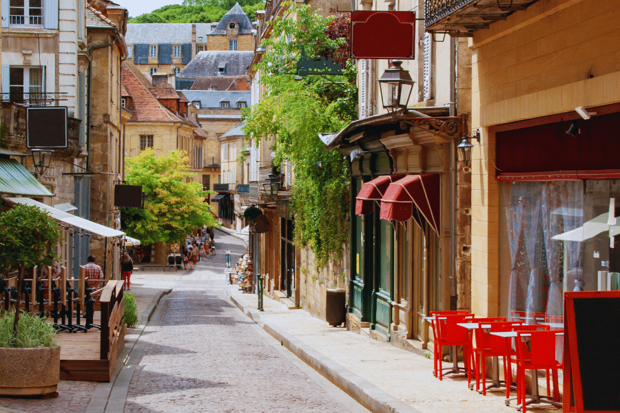 Sarlat-la-Caneda-France A charming narrow street in a European town, flanked by historical buildings with shops and cafes. Small tables with red chairs are set outside a cafe on the right. On the left, people are seated at a shaded outdoor dining area. Green plants adorn the walls.