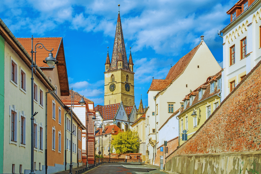 Sibiu Romania A picturesque, narrow street in a European town lined with pastel-colored buildings leads to a prominent church with a tall, intricate steeple. The sky is bright blue with a few clouds, and a stone wall runs alongside the right side of the street.