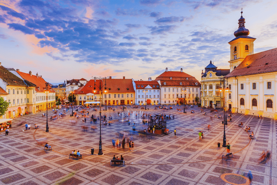 Sibiu Romania A vibrant public square at sunset with a tiled pavement, historic buildings, and a clock tower. People are scattered around, some sitting on benches, while others stroll or gather in groups. The sky is a blend of blue and orange hues, adding a warm ambiance.