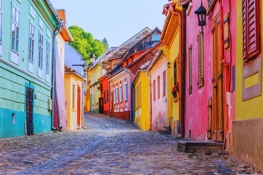 Sighisoara Romania A cobblestone street winds through a vibrant, colorful neighborhood with houses painted in various shades of green, yellow, pink, and red. The buildings exhibit a mix of architectural styles, and the scene is bathed in bright, natural light under a clear blue sky.