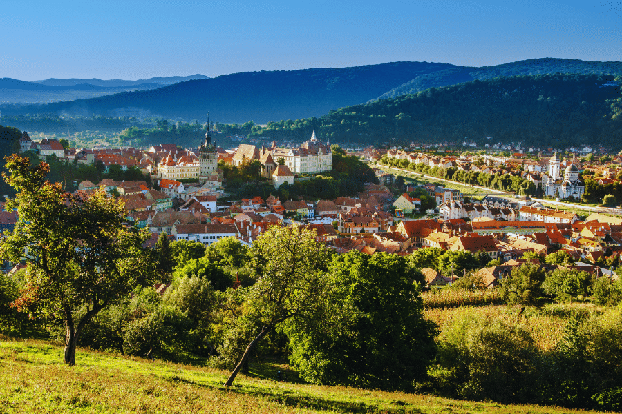 Sighisoara Romania A lush green hillside overlooks a picturesque medieval town with red-tiled rooftops and church steeples nestled in a valley surrounded by forested hills under a clear blue sky. The townscape is bathed in warm sunlight, highlighting its historical charm.