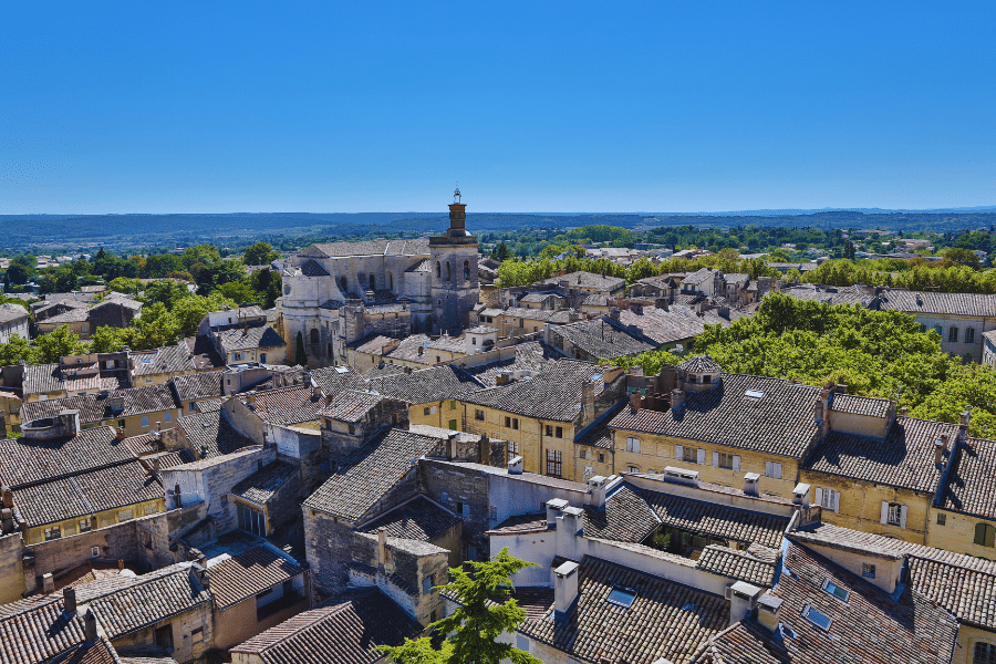 Uzes France An aerial view of a historic town with stone buildings and tiled roofs, centered around a prominent church with a tall tower. Green trees are interspersed among the structures, and rolling hills stretch out into the distance under a clear blue sky.