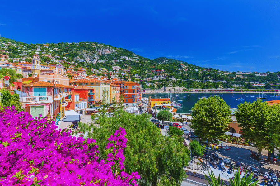 Villefranche-sur-Mer-France A vibrant coastal town features colorful buildings and lush greenery against a backdrop of hills and a blue sky. Purple flowers in the foreground overlook a lively harbor with boats docked in the clear blue water.